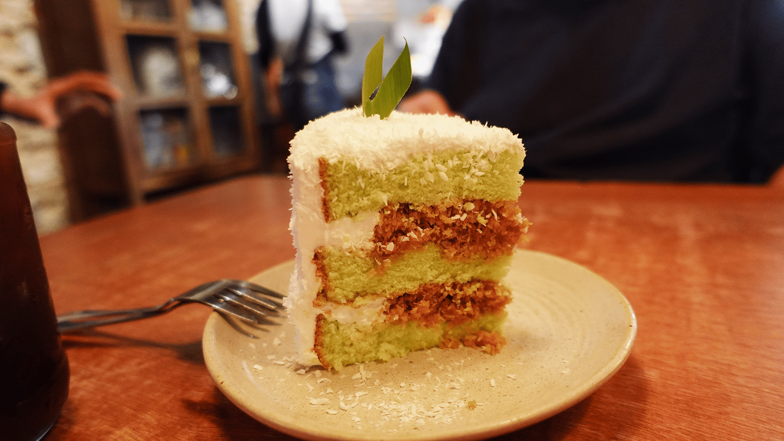 A slice of ondeh-ondeh cake at The Daily Fix Melaka, topped with fragrant pandan, grated coconut, and gula Melaka, a classic melaka food dessert served on a wooden plate.